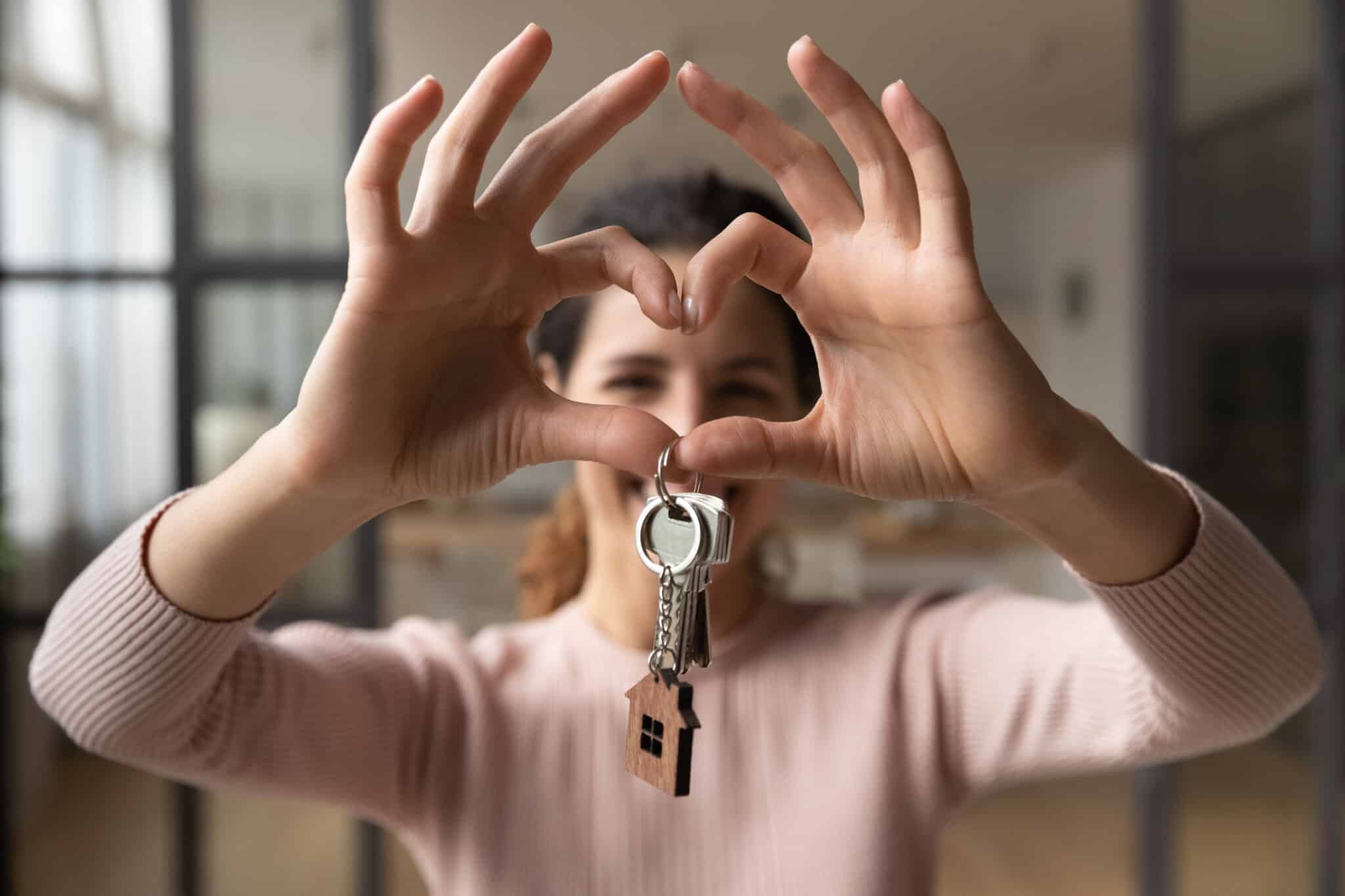 Happy woman hold keys makes heart symbol with joined fingers